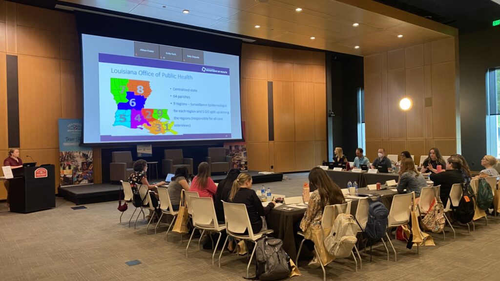 A conference room with attendees seated at tables facing a large presentation screen. The screen displays a slide from the Louisiana Office of Public Health showing a color‑coded map of regional divisions and bullet‑point information. A presenter stands at a podium on the left, while participants take notes and work on laptops. The room has wood-paneled walls, modern lighting, and large windows on the right.